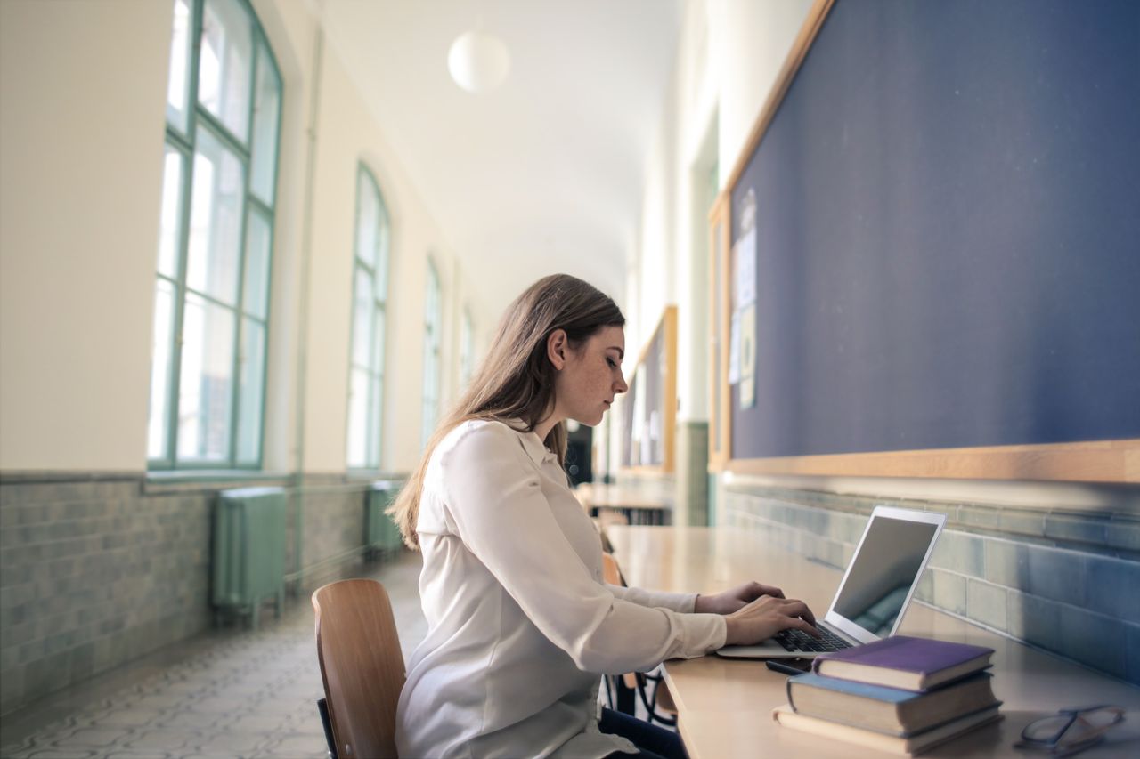 A woman in white blouse working on a laptop in a bright school hallway.